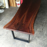 A live-edge wooden table with a rich dark reddish-brown grain pattern, supported by a black metal U-shaped base, photographed in a workshop setting on a concrete floor.
