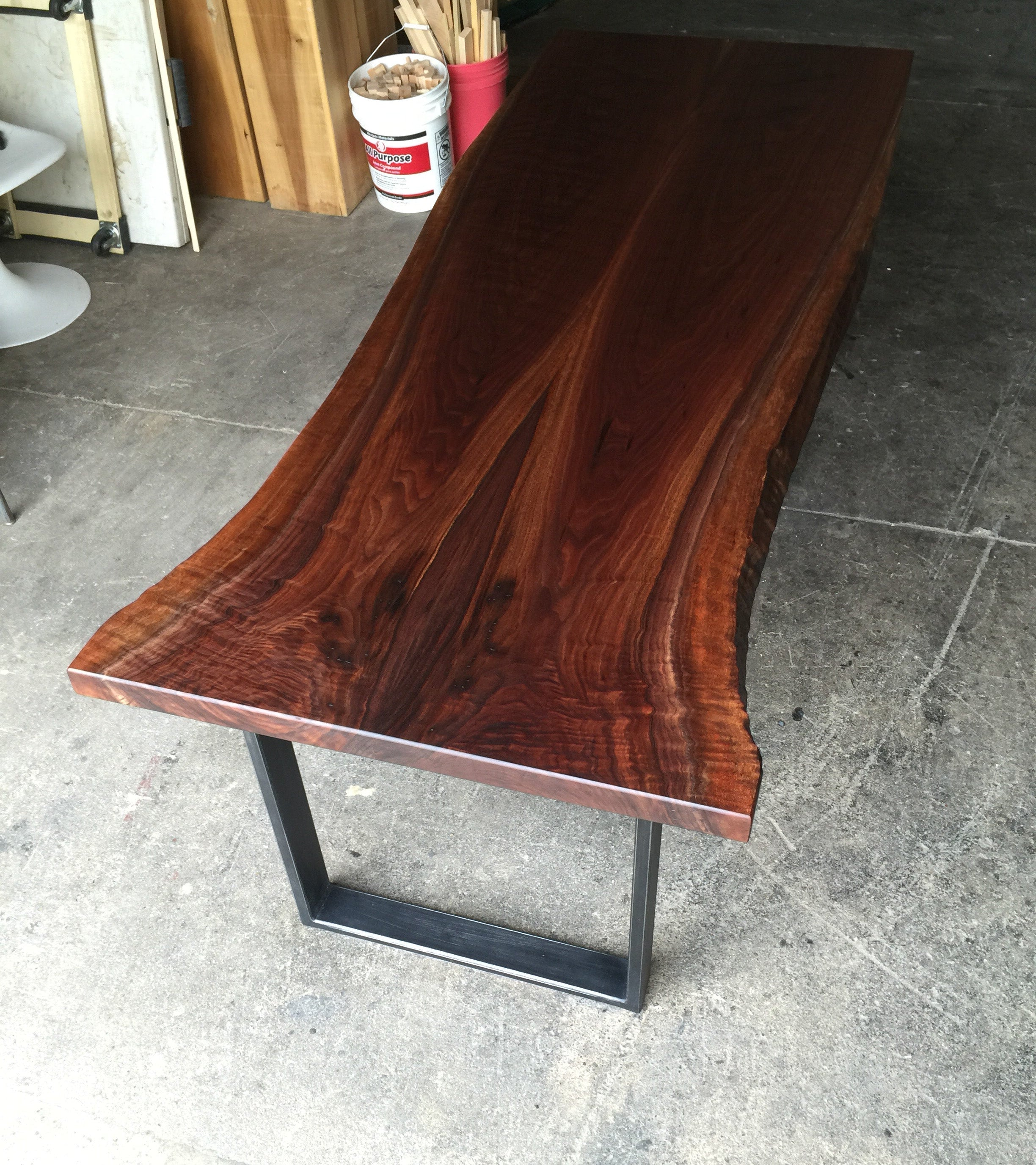 A live-edge wooden table with a rich dark reddish-brown grain pattern, supported by a black metal U-shaped base, photographed in a workshop setting on a concrete floor.