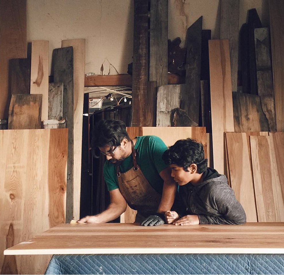 Two men working in a wood workshop, with one wearing an apron and the other assisting, surrounded by large stacks of raw wood planks.
