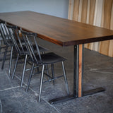 A long rectangular wooden dining table with a dark brown wood top and industrial metal T-leg base, accompanied by four black metal chairs with spindle backs, set in an industrial room with a stone floor and wood-paneled wall.