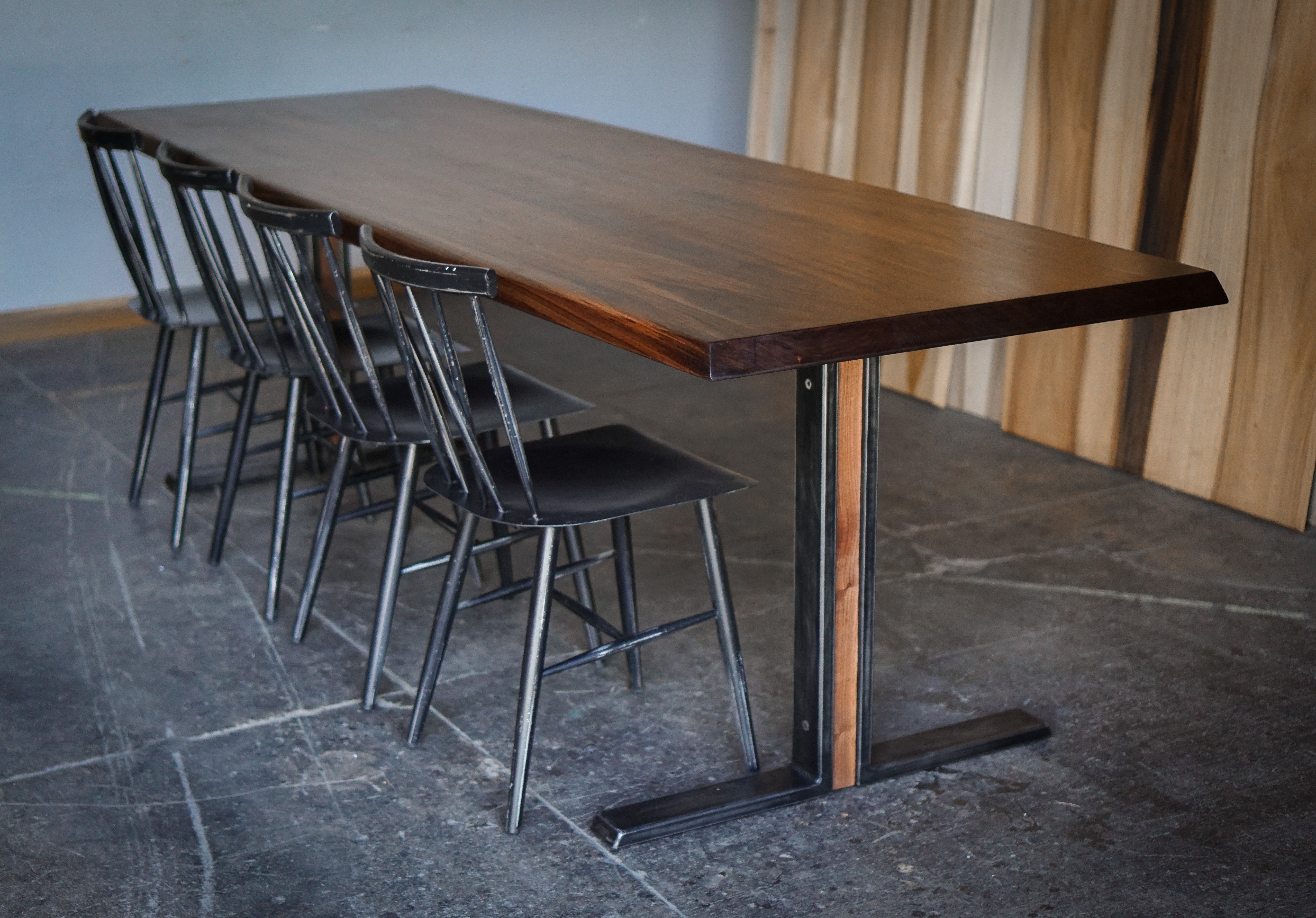 A long rectangular wooden dining table with a dark brown wood top and industrial metal T-leg base, accompanied by four black metal chairs with spindle backs, set in an industrial room with a stone floor and wood-paneled wall.
