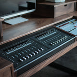 A close-up of a dark wood desk featuring a large black audio mixing console and a smaller control panel integrated into the desk surface.