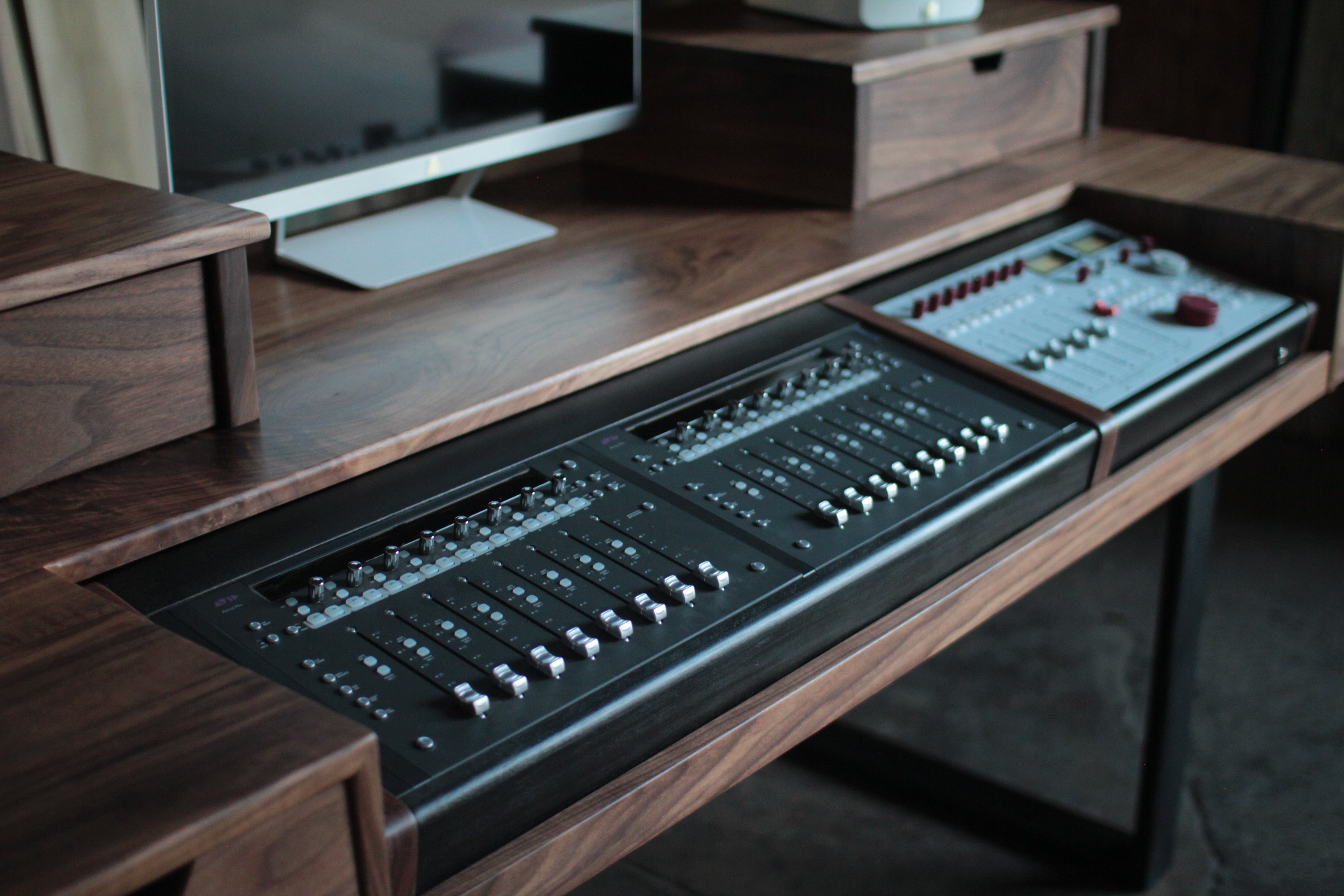 A close-up of a dark wood desk featuring a large black audio mixing console and a smaller control panel integrated into the desk surface.