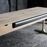 A modern light oak wood console table with a T-shaped brass leg, featuring a drawer on the left and a pull-out shelf on the right holding a black rectangular item.