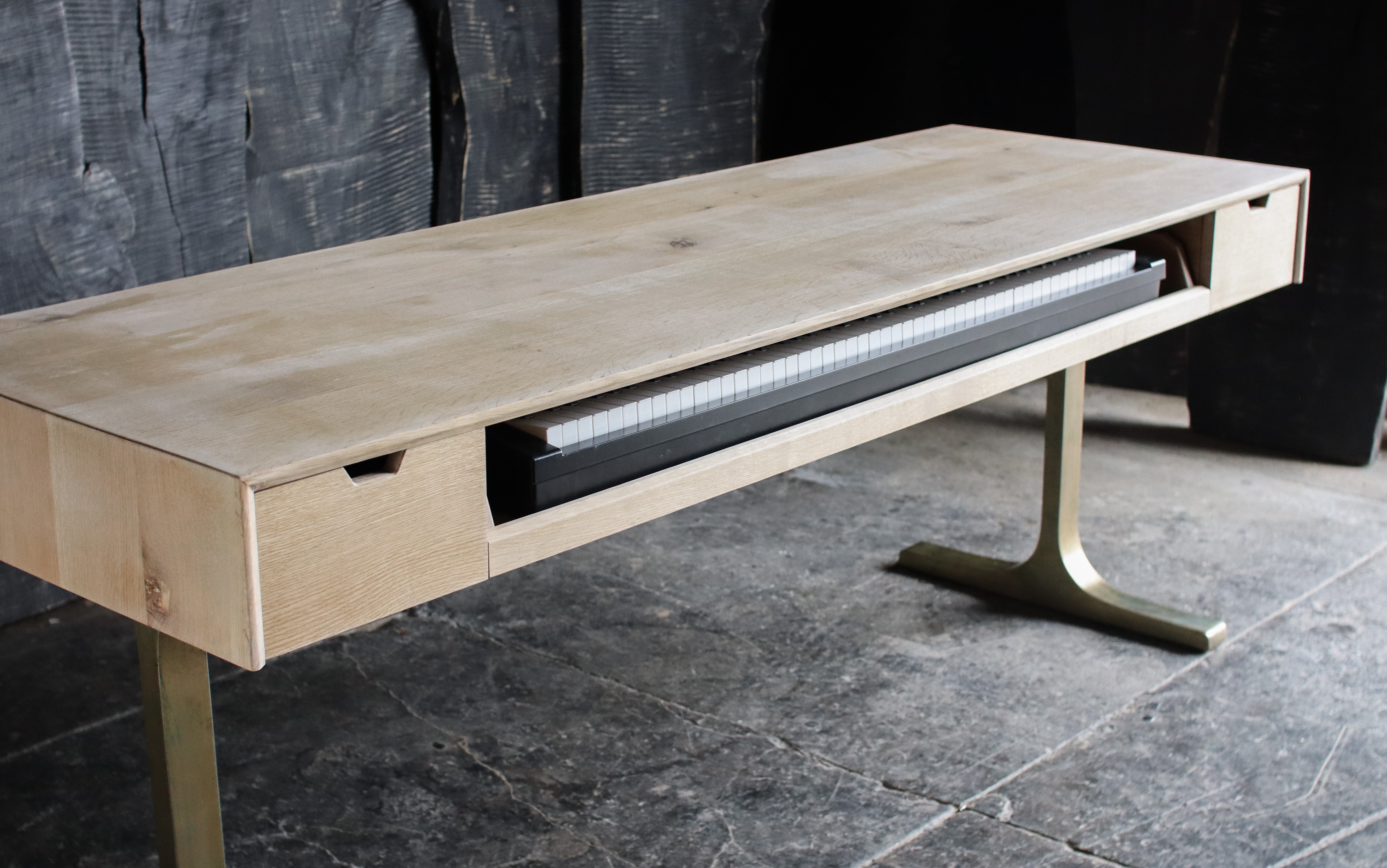 A modern light oak wood console table with a T-shaped brass leg, featuring a drawer on the left and a pull-out shelf on the right holding a black rectangular item.