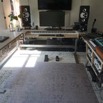 A home studio setup featuring a large wooden height-adjustable desk on the left, a keyboard on a stand in the center, two black speakers, and audio mixing equipment on wooden racks to the right, all set on a patterned rug in a room with bright window light.