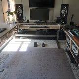 A home studio setup featuring a large wooden height-adjustable desk on the left, a keyboard on a stand in the center, two black speakers, and audio mixing equipment on wooden racks to the right, all set on a patterned rug in a room with bright window light.