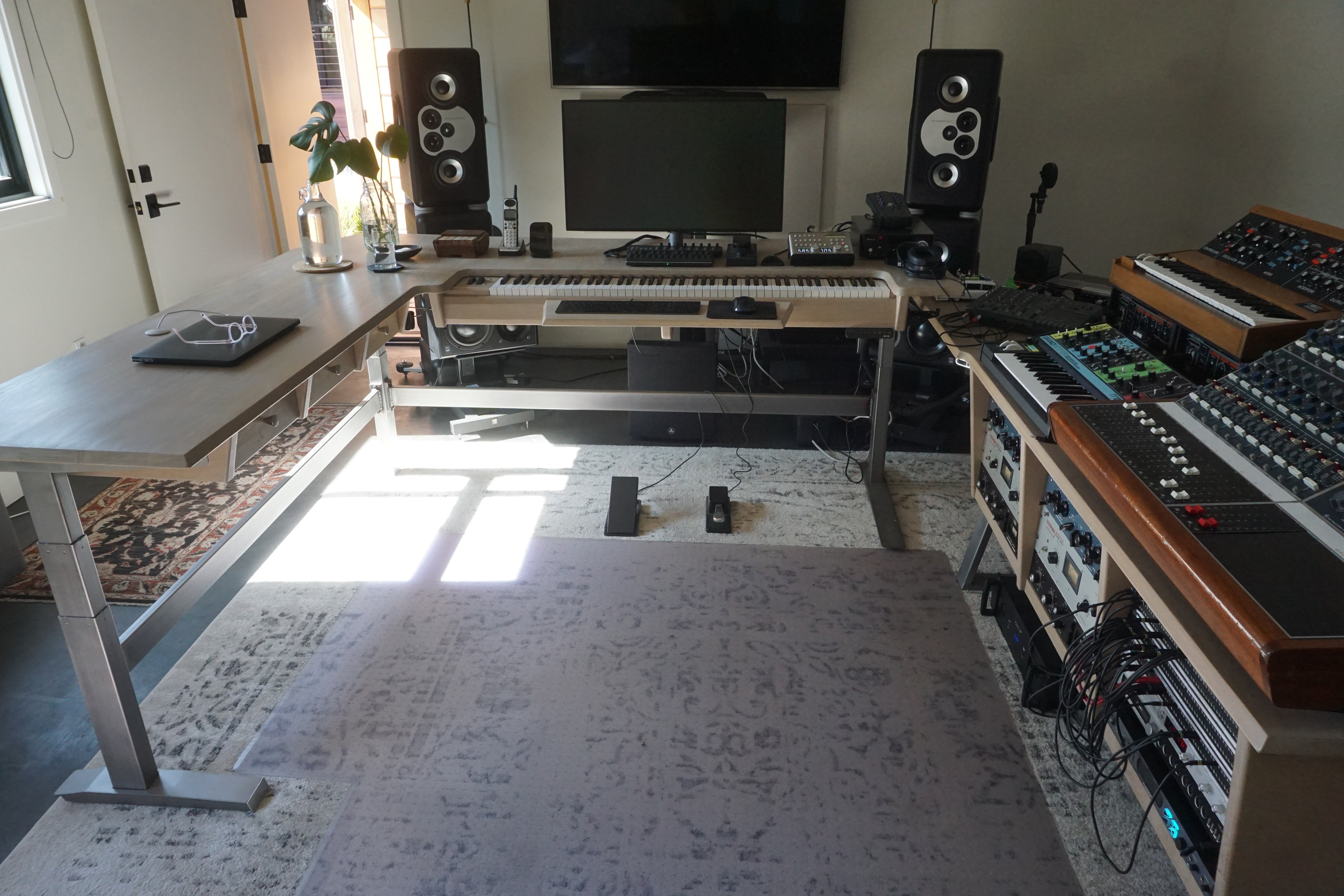 A home studio setup featuring a large wooden height-adjustable desk on the left, a keyboard on a stand in the center, two black speakers, and audio mixing equipment on wooden racks to the right, all set on a patterned rug in a room with bright window light.