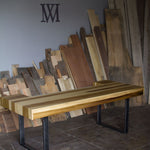 A rustic wooden dining table with a striped pattern of light and dark wood planks, supported by black metal U-shaped legs, set in a workshop with wood scraps stacked in the background.