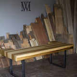 A rustic wooden dining table with a striped pattern of light and dark wood planks, supported by black metal U-shaped legs, set in a workshop with wood scraps stacked in the background.