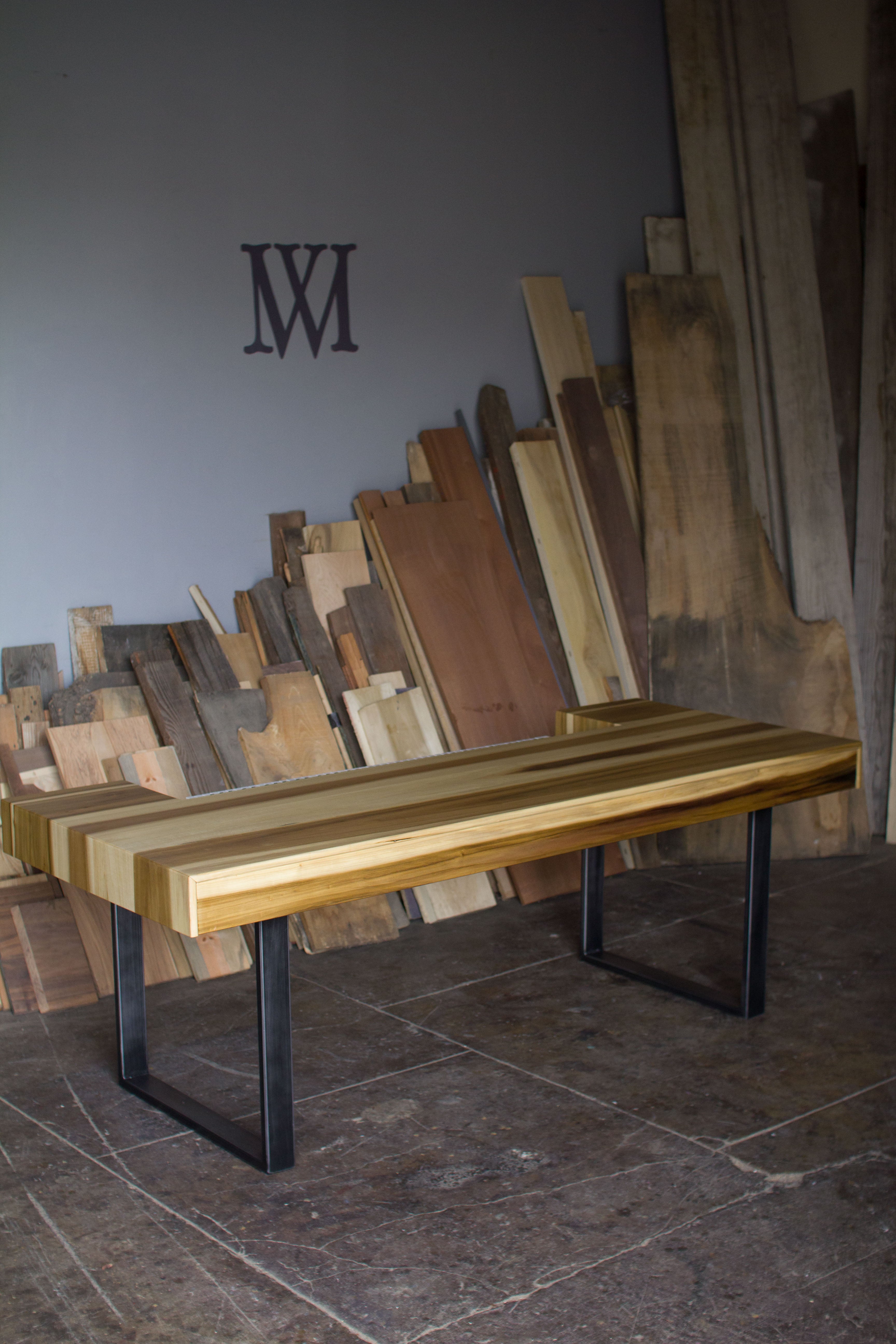 A rustic wooden dining table with a striped pattern of light and dark wood planks, supported by black metal U-shaped legs, set in a workshop with wood scraps stacked in the background.