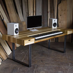 A rustic computer desk made of light-colored wood with black metal legs, styled with a computer monitor, keyboard, and two white speakers against a background of vertical wooden planks.