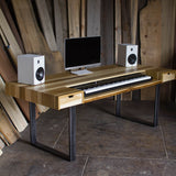 A rustic computer desk made of light-colored wood with black metal legs, styled with a computer monitor, keyboard, and two white speakers against a background of vertical wooden planks.