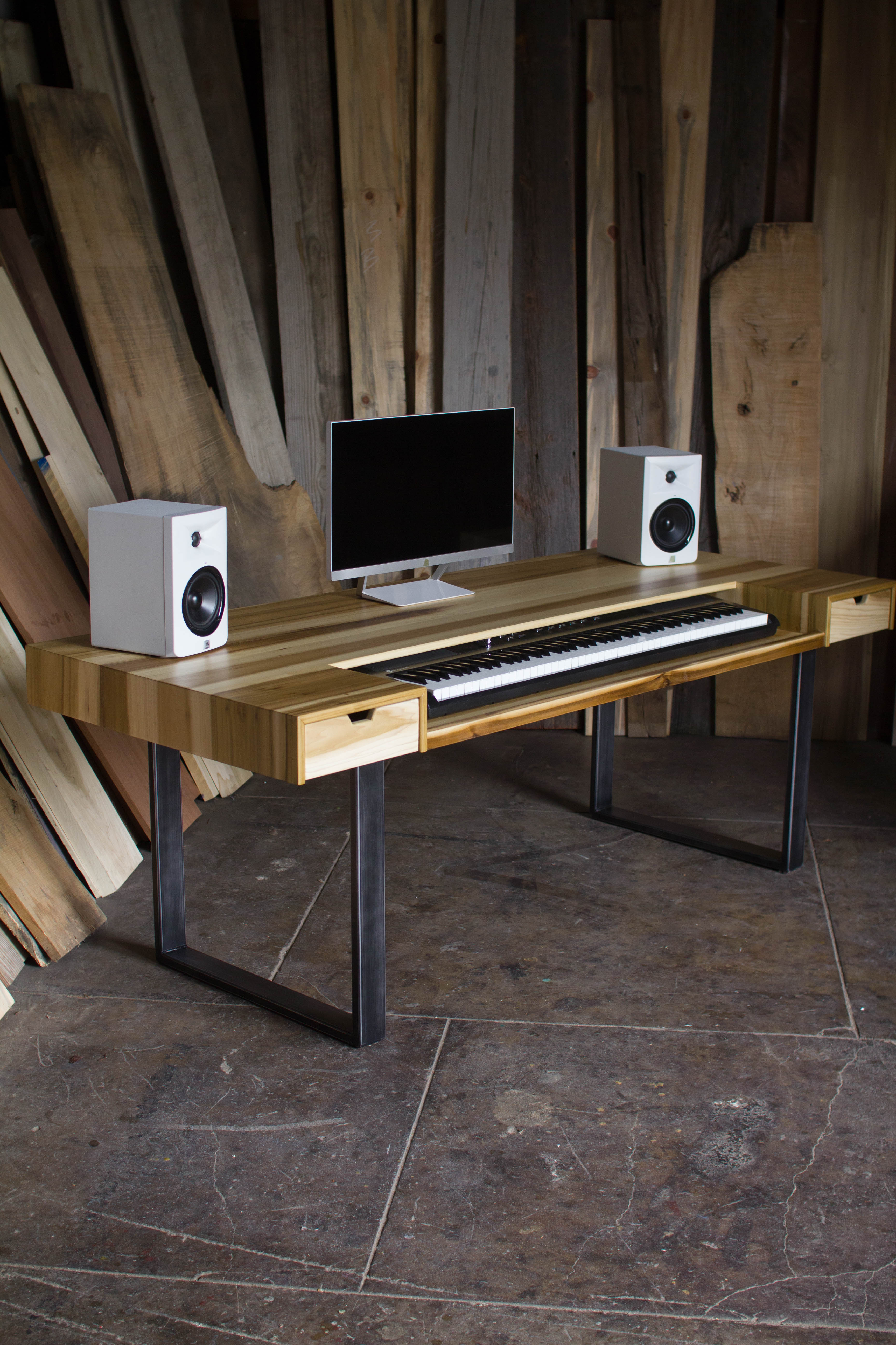 A rustic computer desk made of light-colored wood with black metal legs, styled with a computer monitor, keyboard, and two white speakers against a background of vertical wooden planks.