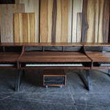 A large wooden multi-keyboard workstation or piano console with angled metal legs, shown in a workshop-like interior with wood panels on the wall and a dark floor.