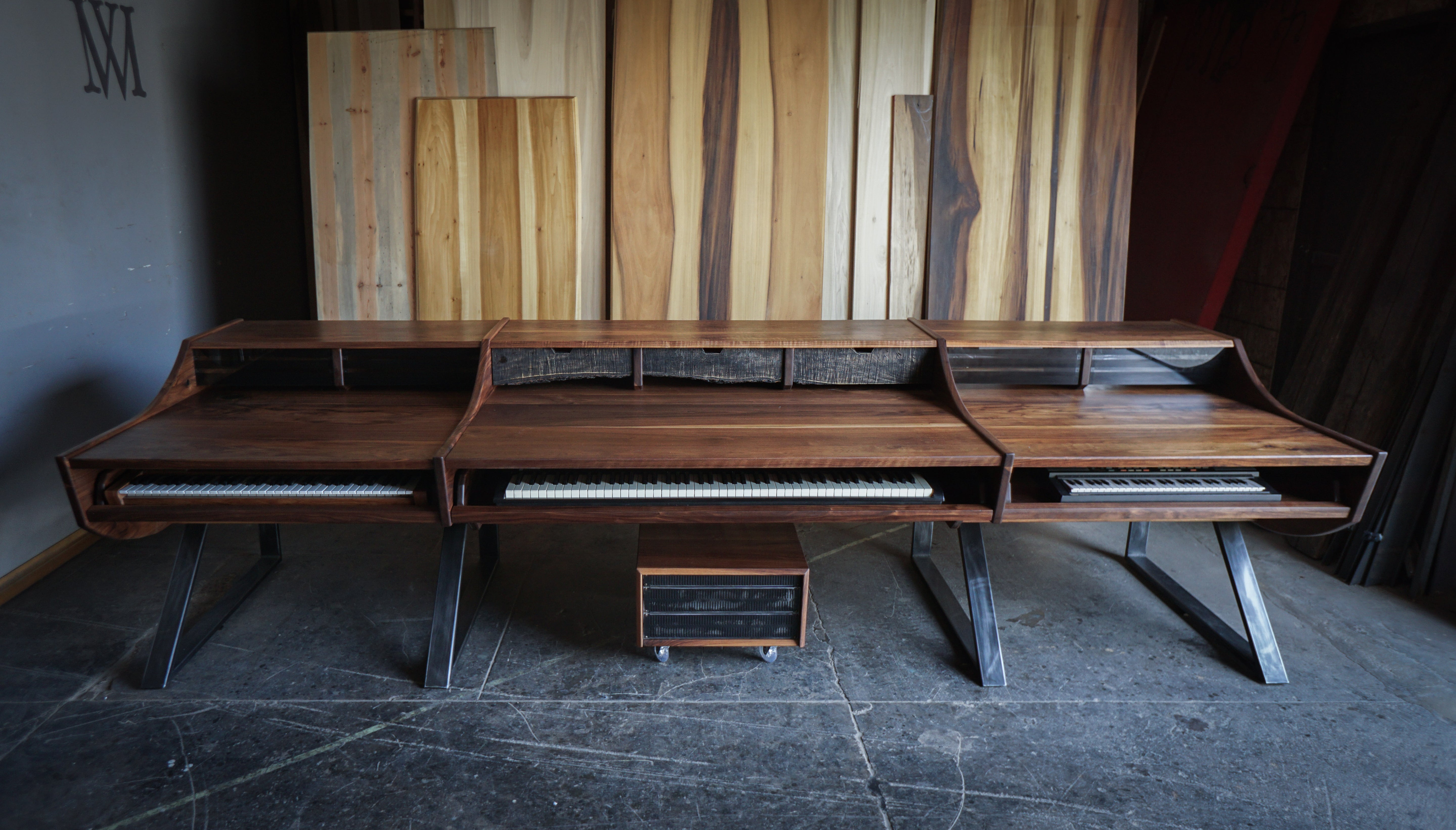 A large wooden multi-keyboard workstation or piano console with angled metal legs, shown in a workshop-like interior with wood panels on the wall and a dark floor.