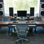 A large custom-built wooden music production desk in a recording studio, featuring a keyboard, MIDI controller, computer monitor, speakers, and an office chair in front.