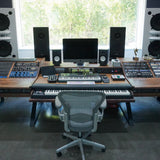 A large custom-built wooden music production desk in a recording studio, featuring a keyboard, MIDI controller, computer monitor, speakers, and an office chair in front.