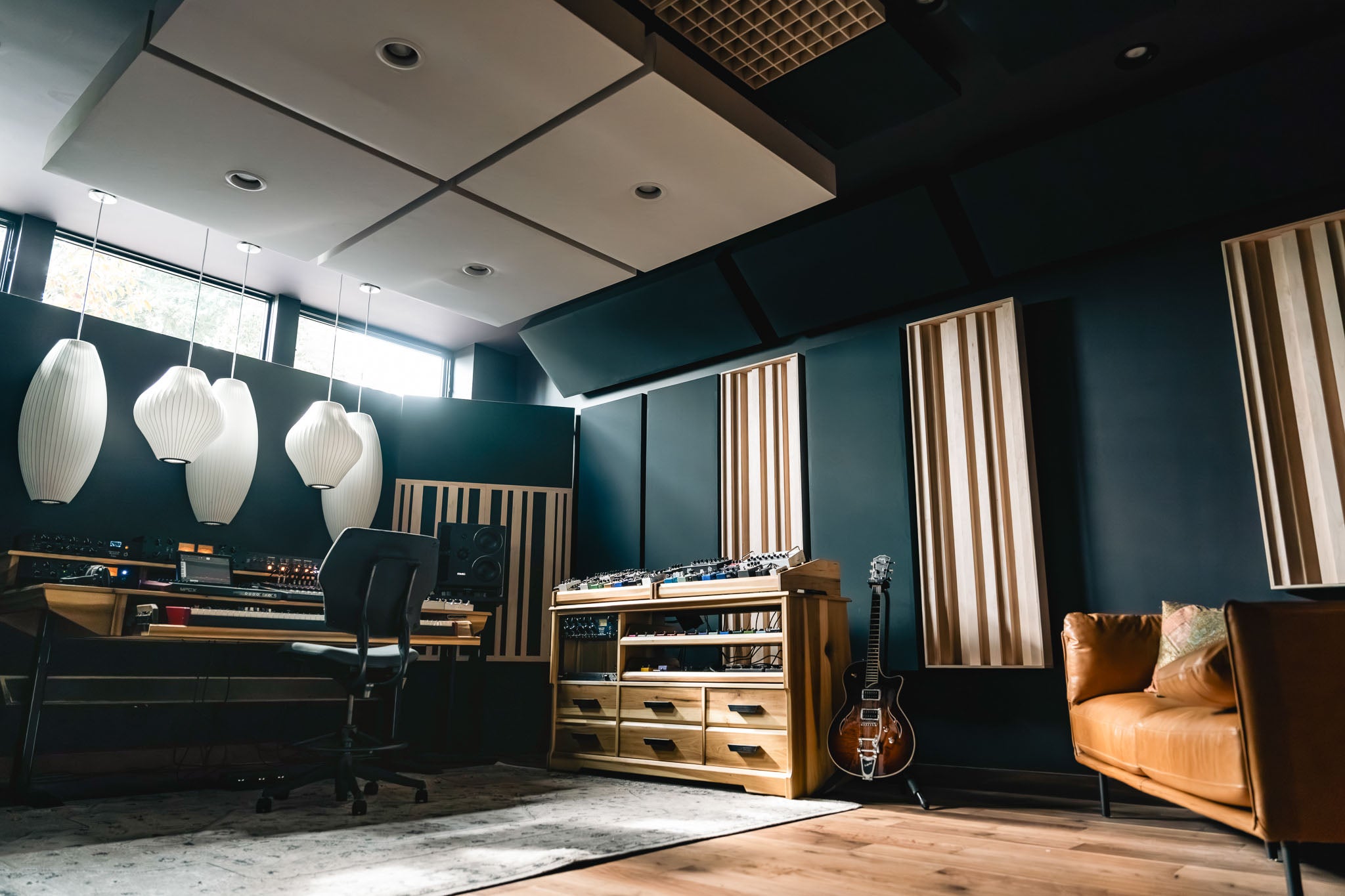 A modern music studio interior with dark blue walls, wood acoustic panels, pendant lights, a mixing desk, a wooden storage cabinet, a guitar, and a tan leather sofa.