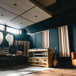 A modern music studio interior with dark blue walls, wood acoustic panels, pendant lights, a mixing desk, a wooden storage cabinet, a guitar, and a tan leather sofa.