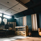 A modern music studio interior with dark blue walls, wood acoustic panels, pendant lights, a mixing desk, a wooden storage cabinet, a guitar, and a tan leather sofa.