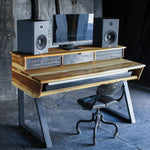 A rustic, industrial-style computer desk made from reclaimed wood and scrap metal, styled with a computer monitor, two speakers, and a black office chair in a dark, moody room.