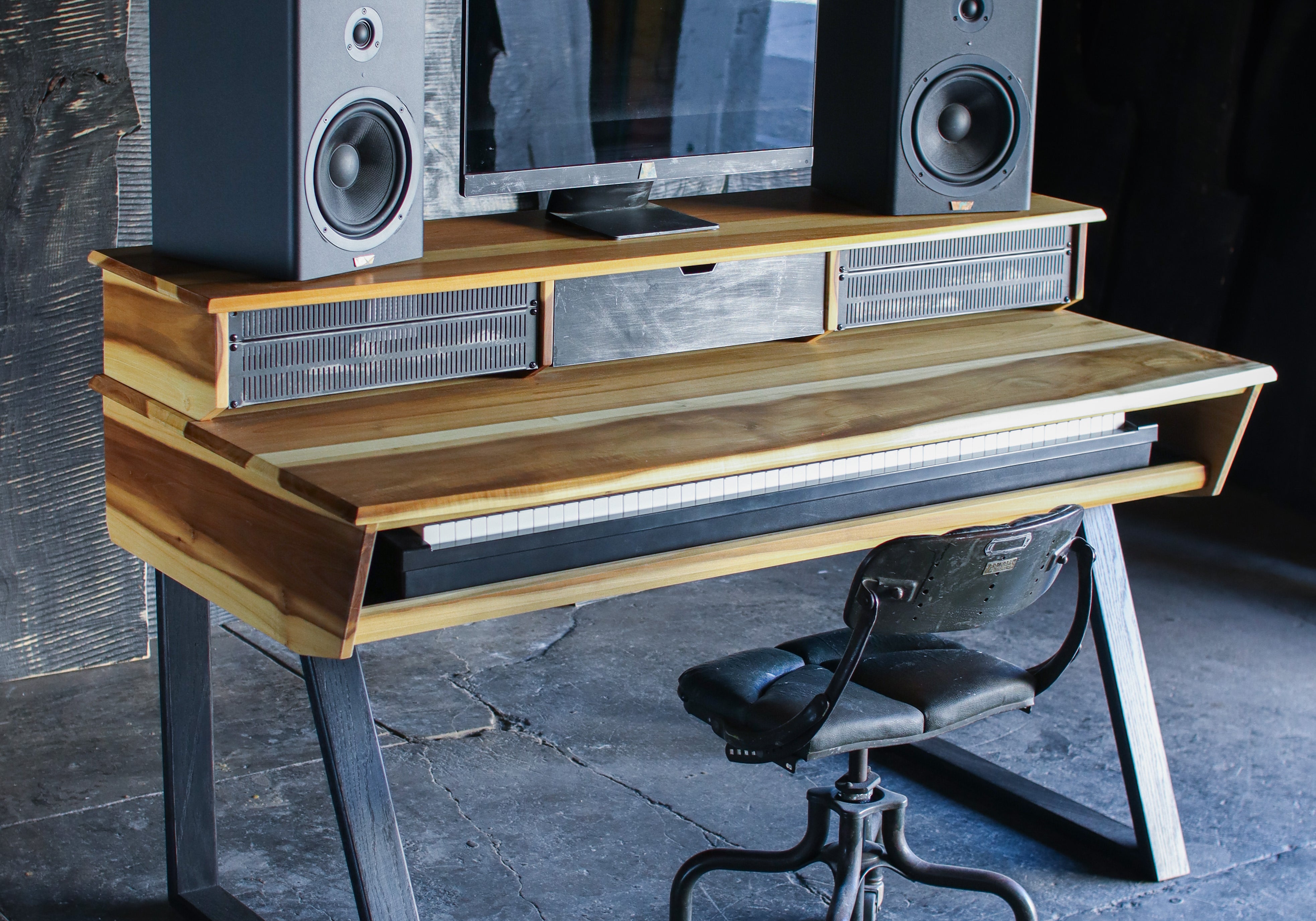 A rustic, industrial-style computer desk made from reclaimed wood and scrap metal, styled with a computer monitor, two speakers, and a black office chair in a dark, moody room.