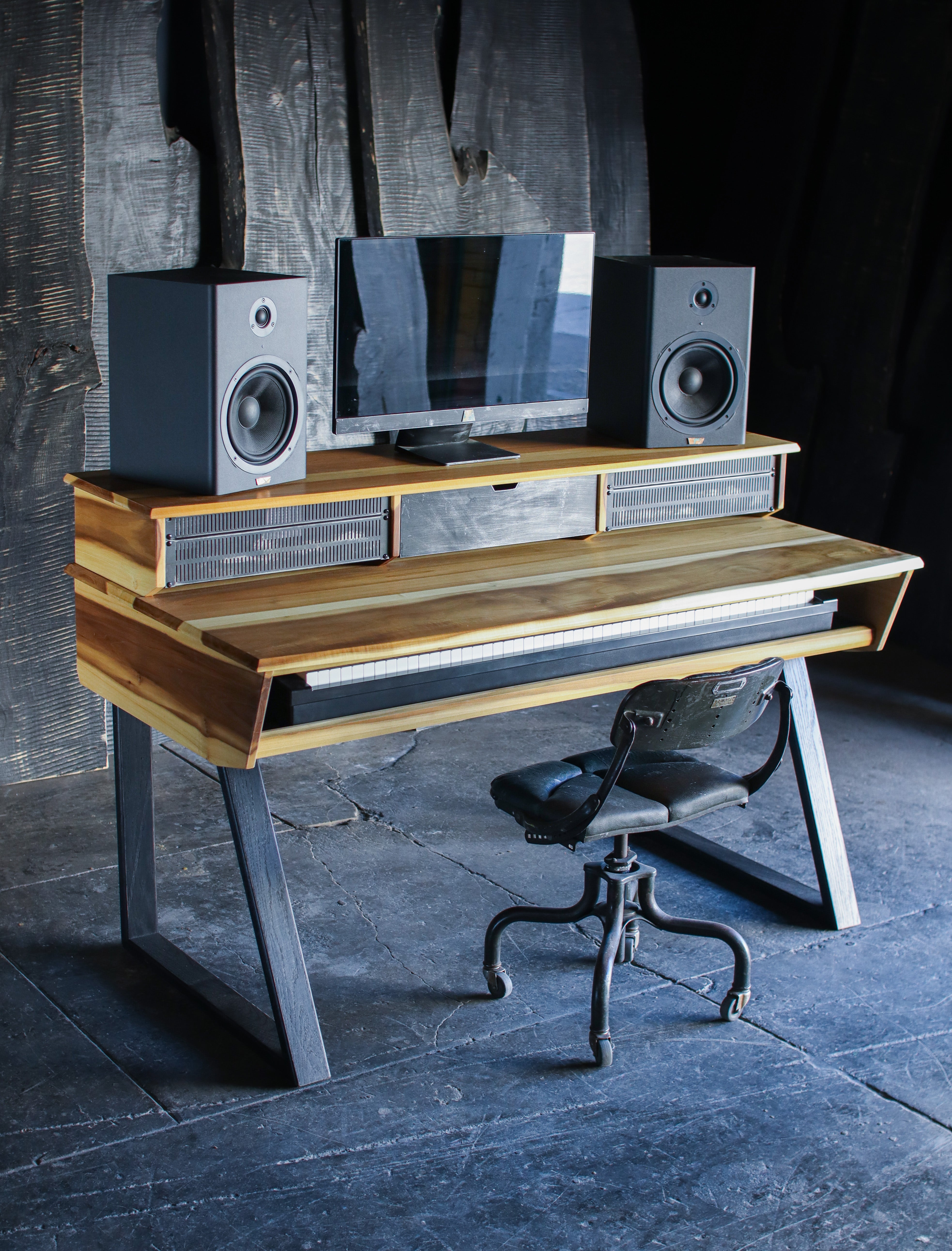 A rustic, industrial-style computer desk made from reclaimed wood and scrap metal, styled with a computer monitor, two speakers, and a black office chair in a dark, moody room.
