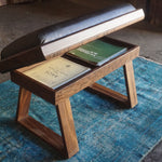 A wooden bench with a black cushioned top and open storage compartment, placed on a blue rug in a rustic workshop setting.