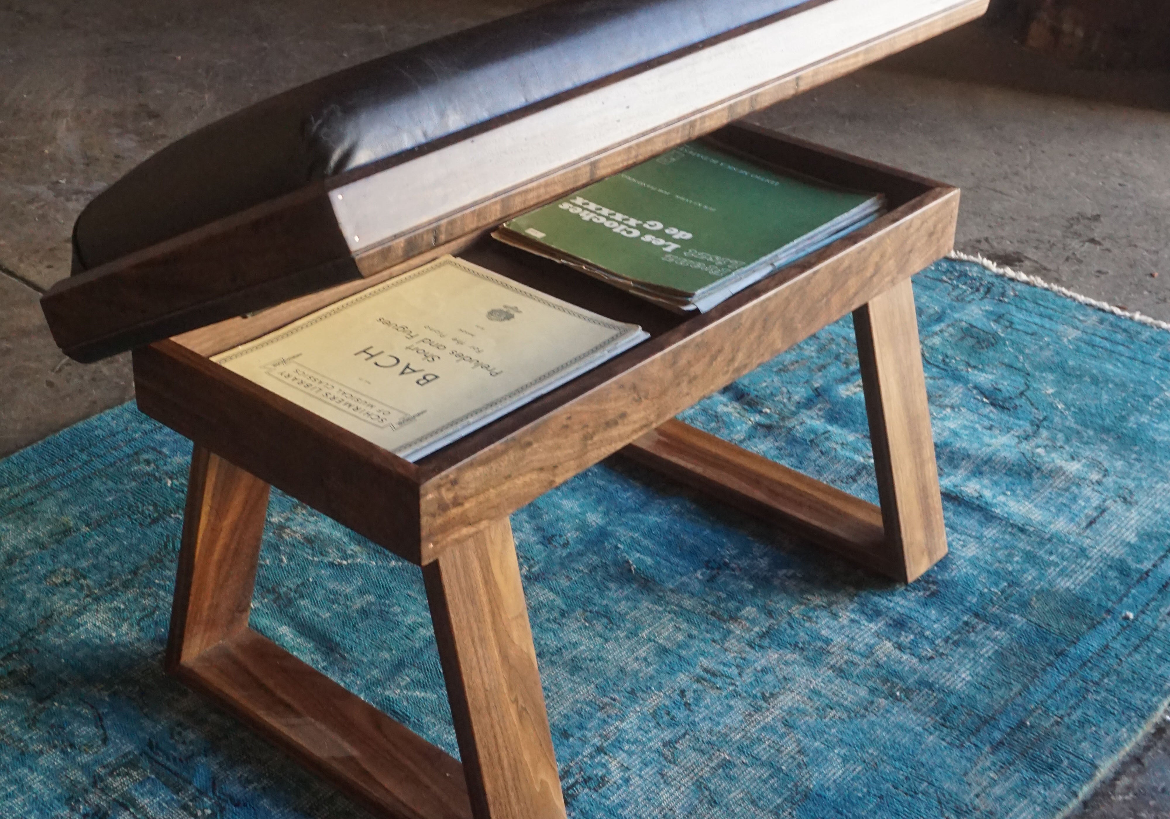 A wooden bench with a black cushioned top and open storage compartment, placed on a blue rug in a rustic workshop setting.