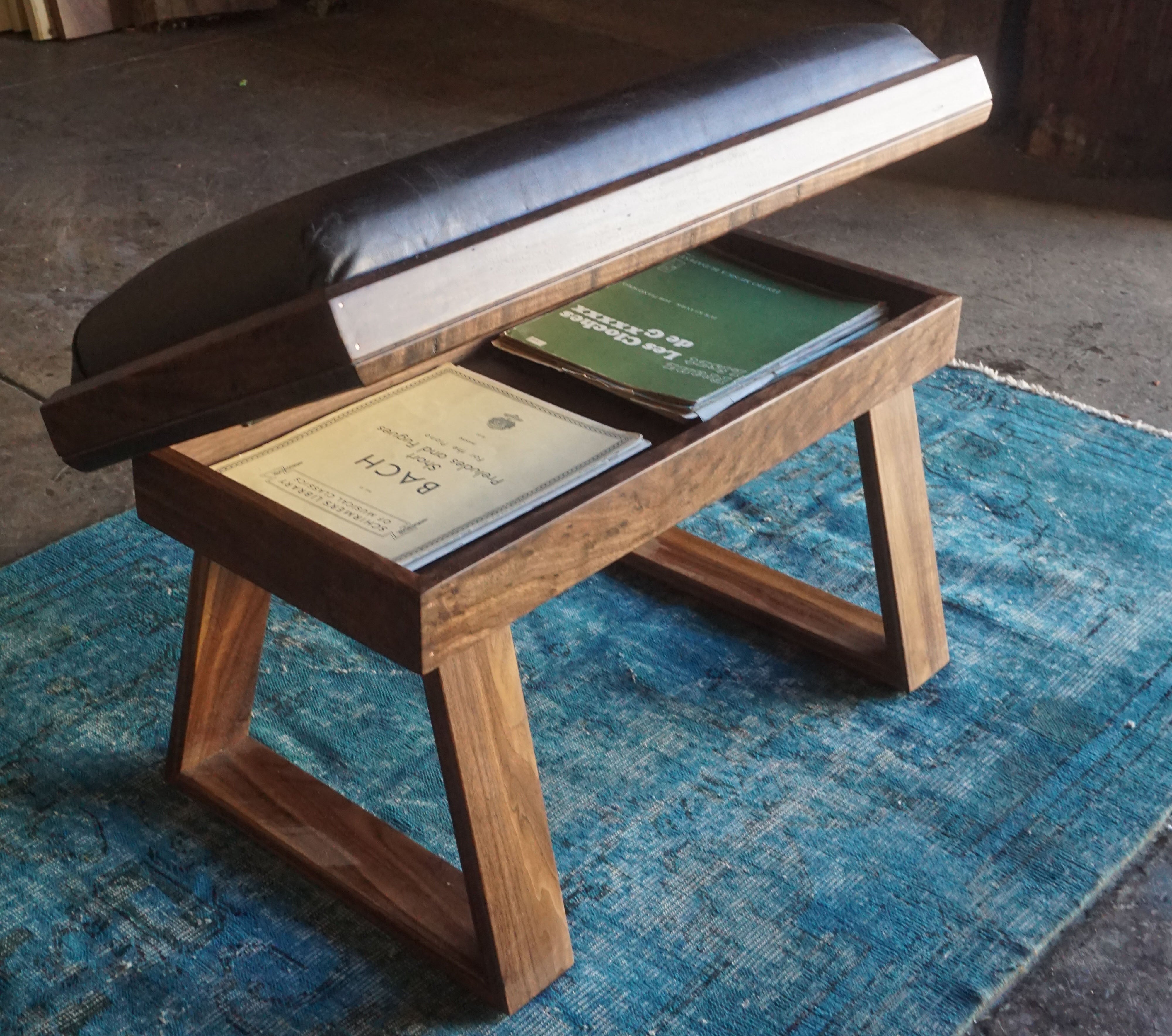 A wooden bench with a black cushioned top and open storage compartment, placed on a blue rug in a rustic workshop setting.