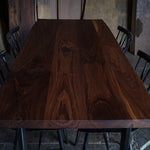 A rectangular dark brown wooden dining table with a rich wood grain finish, surrounded by black metal chairs in a rustic workshop setting.