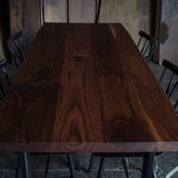 A rectangular dark brown wooden dining table with a rich wood grain finish, surrounded by black metal chairs in a rustic workshop setting.