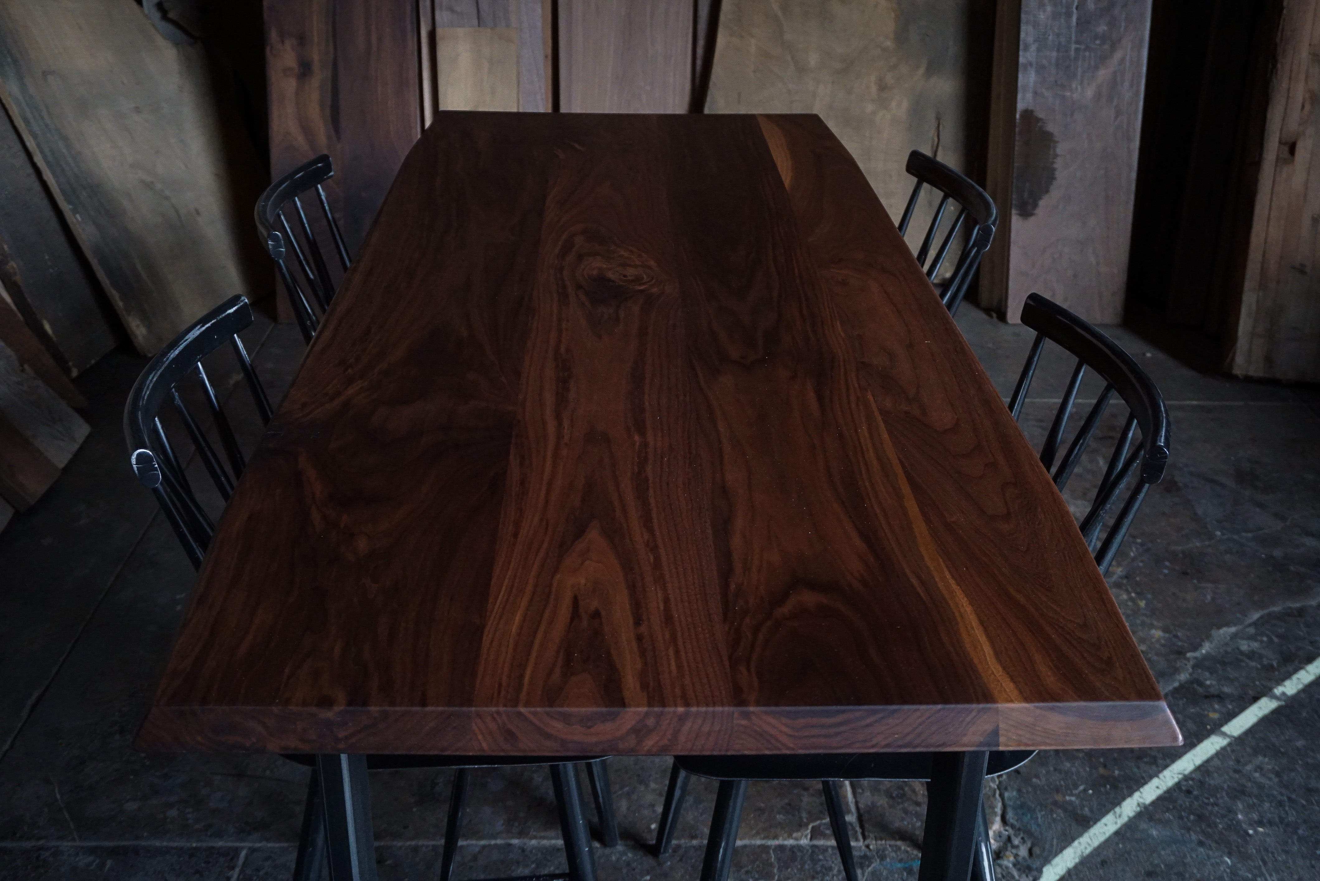 A rectangular dark brown wooden dining table with a rich wood grain finish, surrounded by black metal chairs in a rustic workshop setting.