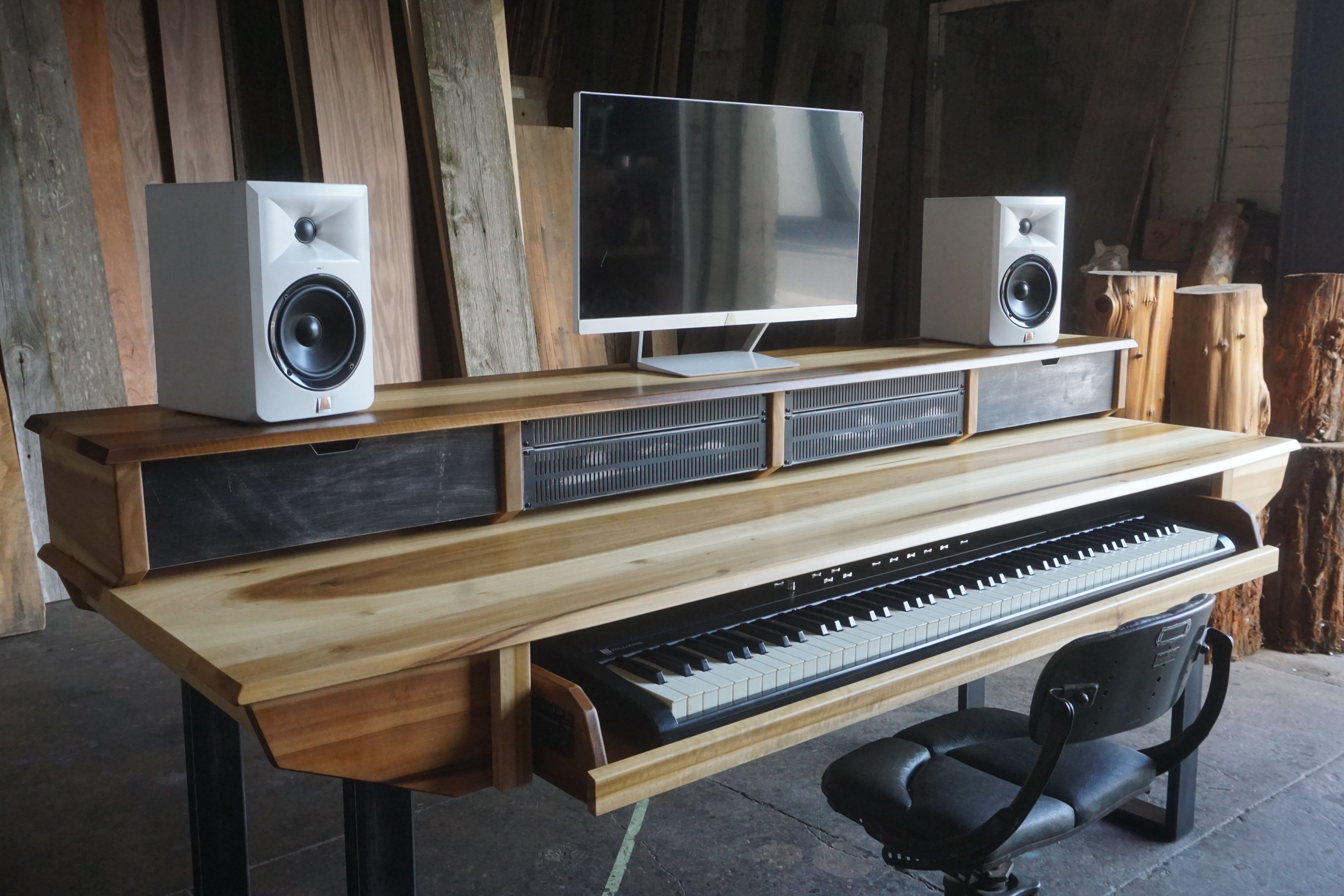 Wooden studio desk with electronic keyboard, computer monitor, and speakers in a rustic room.