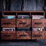 A rustic wooden record storage cabinet with nine drawers, some open to reveal vinyl records inside, set against a dark wood plank background.
