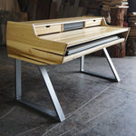 A modern wooden writing desk with a light natural wood finish and silver metal legs, set in a workshop environment with wood panels and books in the background.