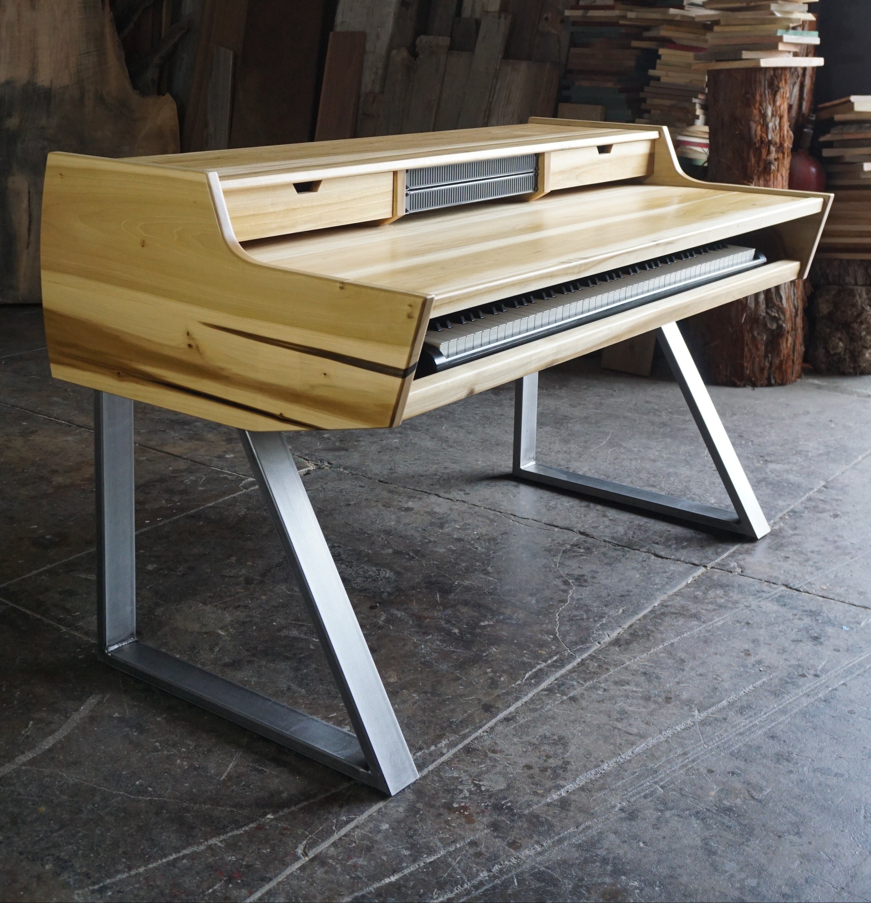 A modern wooden writing desk with a light natural wood finish and silver metal legs, set in a workshop environment with wood panels and books in the background.