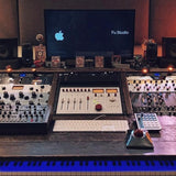 A large audio mixing console setup on a wooden desk in a studio environment, with a central monitor displaying the Apple logo and the text 'Fu Studio', flanked by speakers, multiple control modules, a keyboard, and a piano keyboard visible at the bottom.