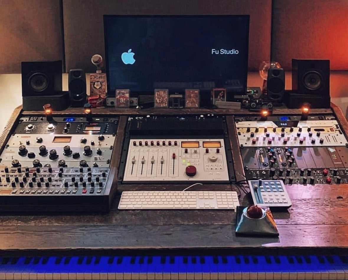 A large audio mixing console setup on a wooden desk in a studio environment, with a central monitor displaying the Apple logo and the text 'Fu Studio', flanked by speakers, multiple control modules, a keyboard, and a piano keyboard visible at the bottom.